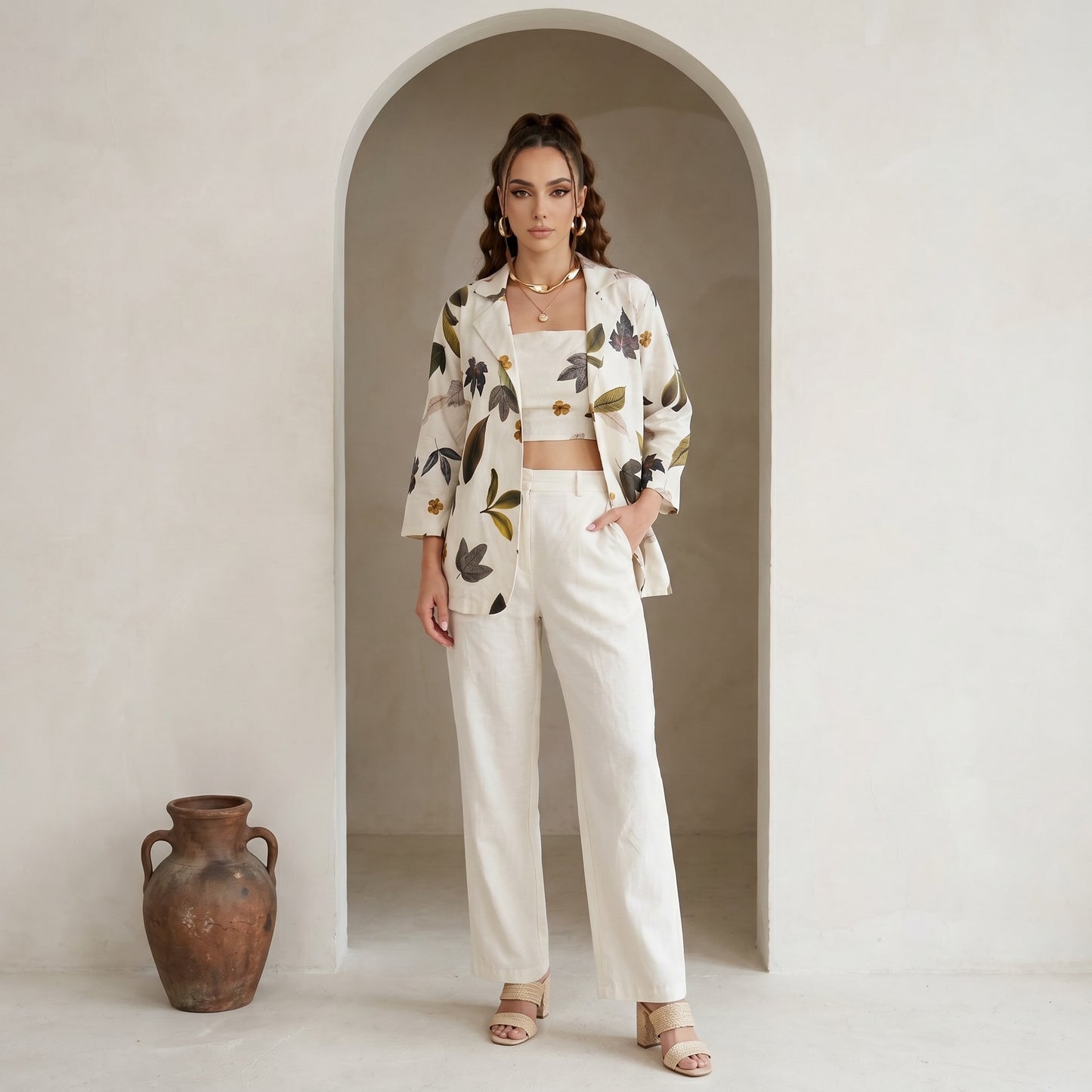 Woman in a floral jacket and white pants standing in an archway with a vase on the floor.
