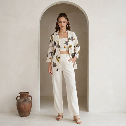 Woman in a floral jacket and white pants standing in an archway with a vase on the floor.
